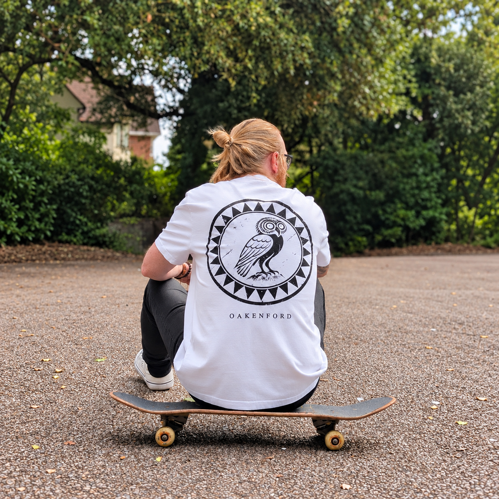 Seated model wearing the white Owl Shield Oakenford graphic T-shirt on a skateboard