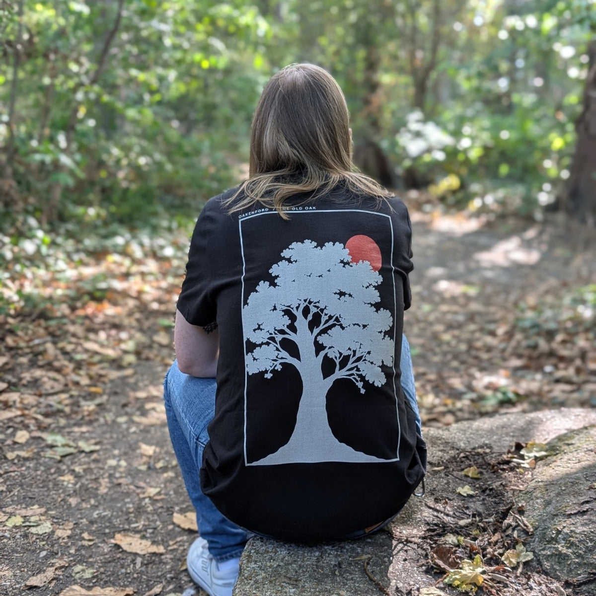 Seated model wearing The Old Oak black T-shirt, back view in woodland